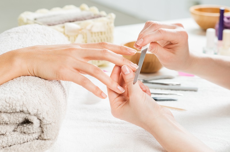 Manicure w Rzeszowie  Na zdjęciu widzimy delikatne dłonie klientki, której paznokcie są starannie pielęgnowane przez kosmetyczkę. Kosmetyczka używa pilnika do nadania paznokciom odpowiedniego kształtu. Na stole widać różnorodne narzędzia manicure oraz kosmetyki, które służą do wykonania zabiegu. Wszystko to odbywa się w przyjemnej, relaksującej atmosferze, co podkreśla dbałość o szczegóły i profesjonalizm salonu kosmetycznego w Rzeszowie.