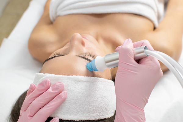 Na zdjęciu widoczna jest kobieta poddawana zabiegowi oczyszczania wodorowego twarzy. Pacjentka leży na plecach, z opaską na głowie, a kosmetyczka w różowych rękawiczkach delikatnie aplikuje urządzenie na jej czoło. Oczyszczanie wodorowe jest nowoczesnym zabiegiem, który głęboko oczyszcza skórę, usuwa wolne rodniki i poprawia jej nawilżenie, pozostawiając ją świeżą i promienną.