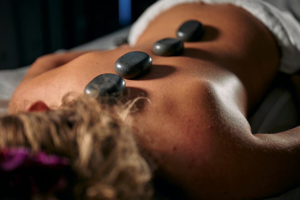 Masaż gorącymi kamieniami - Na zdjęciu widoczna jest osoba leżąca na brzuchu, poddawana masażowi gorącymi kamieniami. Na plecach pacjenta ułożone są trzy ciemne, gładkie kamienie, które mają na celu rozluźnienie mięśni i poprawę krążenia. Osoba jest owinięta ręcznikiem, co sugeruje, że zabieg odbywa się w profesjonalnym salonie spa. Tego typu masaż pomaga w relaksacji, redukcji stresu oraz łagodzeniu bólu mięśniowego.