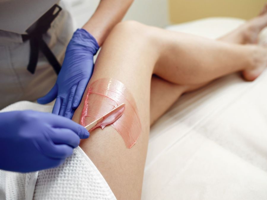różowy wosk nakładany różową szpatułką na udo kobiety przez kobietę w niebieskich gumowych rękawiczkach