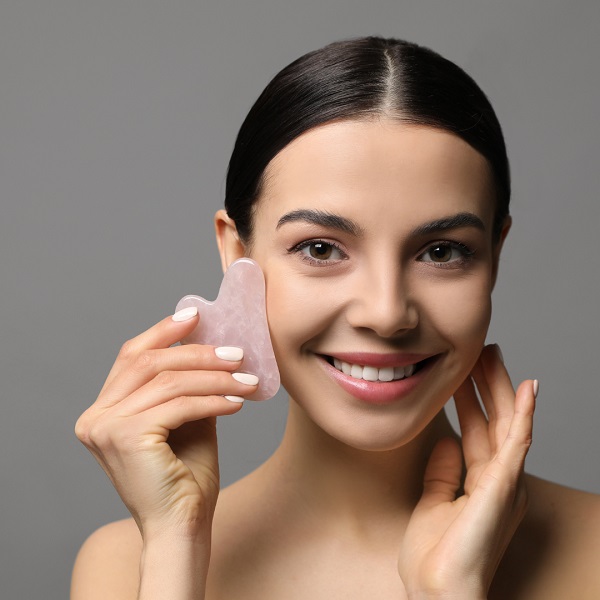 Masaż Gua Sha - Uśmiechnięta kobieta wykonuje masaż twarzy różowym kamieniem Gua Sha, trzymając narzędzie przy policzku – zabieg liftingujący i relaksujący skórę twarzy.