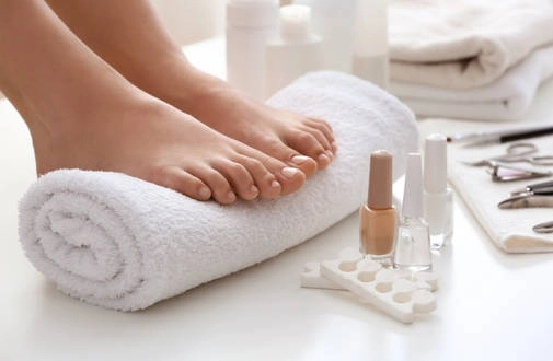 Stopy kobiety opierające się na białym ręczniku, otoczone przyborami do pedicure: lakierami do paznokci, pilnikami i innymi akcesoriami do pielęgnacji stóp.