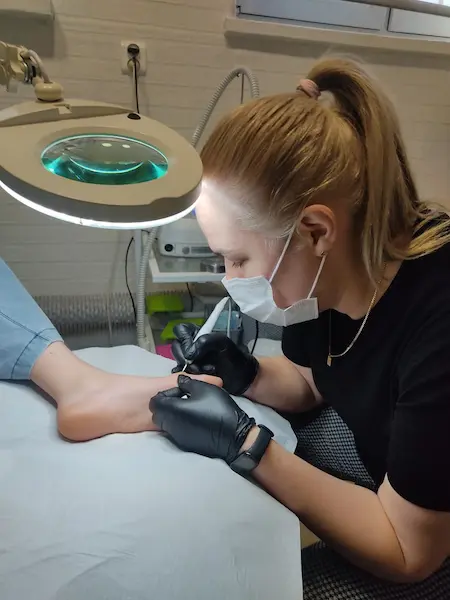 Na zdjęciu widzimy specjalistkę podologa podczas pracy w salonie kosmetycznym. Kobieta, nosząca maseczkę ochronną i rękawiczki, precyzyjnie wykonuje zabieg pielęgnacyjny na stopie klienta. W tle znajduje się lampa powiększająca oraz różne narzędzia potrzebne do zabiegu.