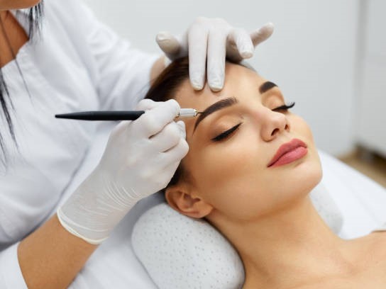 Na zdjęciu widoczny jest zabieg makijażu permanentnego brwi. Pacjentka leży wygodnie na poduszce, mając zamknięte oczy i będąc w stanie relaksu. Specjalista w rękawiczkach precyzyjnie wykonuje makijaż permanentny, używając specjalnego urządzenia. Procedura ta ma na celu trwałe podkreślenie i ukształtowanie brwi, nadając im bardziej zdefiniowany i estetyczny wygląd.