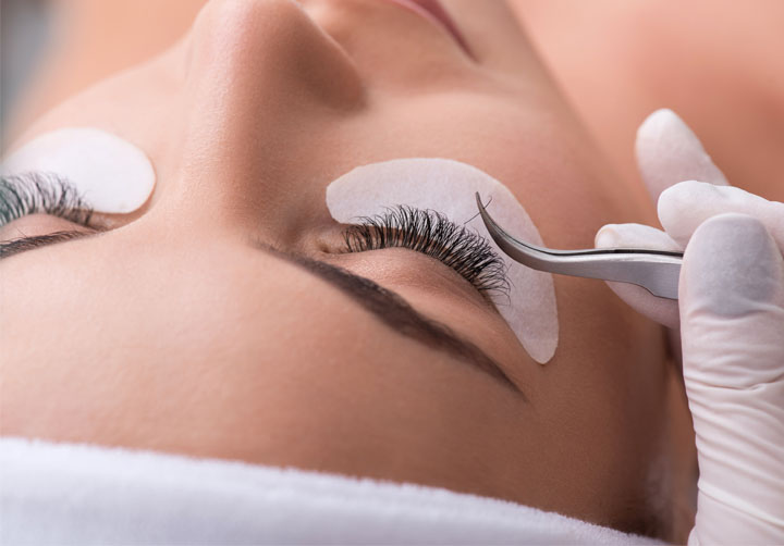 leżaca kobieta z białymi płatkami pod powiekami. druga kobieta w rękawiczkach trzyma pęsetę i przykleja sztuczne rzęsy