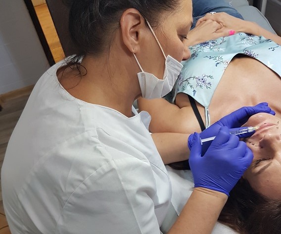 Kosmetologia estetyczna - Zdjęcie przedstawia specjalistkę w białym uniformie medycznym i maseczce, która wykonuje zabieg kosmetologiczny na twarzy leżącej klientki. Specjalistka używa strzykawki do precyzyjnego wstrzyknięcia preparatu, a jej dłonie są zabezpieczone niebieskimi rękawiczkami. Klientka leży wygodnie na plecach, ubrana w bluzkę bez ramiączek. Zdjęcie ilustruje profesjonalizm i dbałość o higienę podczas zabiegu kosmetologii estetycznej.