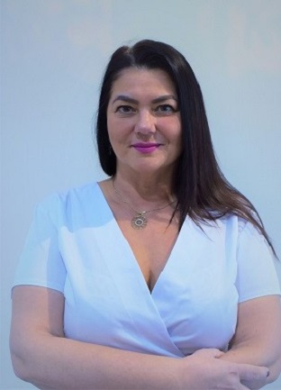 Kobieta z ciemnymi włosami ubrana w biały uniform, stojąca na tle jasnej ściany. Ma na sobie naszyjnik i delikatny makijaż. Uśmiecha się delikatnie, wyglądając profesjonalnie i przyjaźnie.